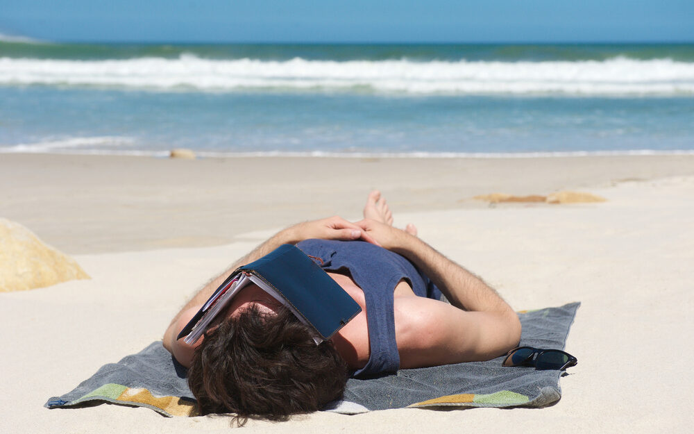 A man sleeping on the beach with a book covering his face for shade